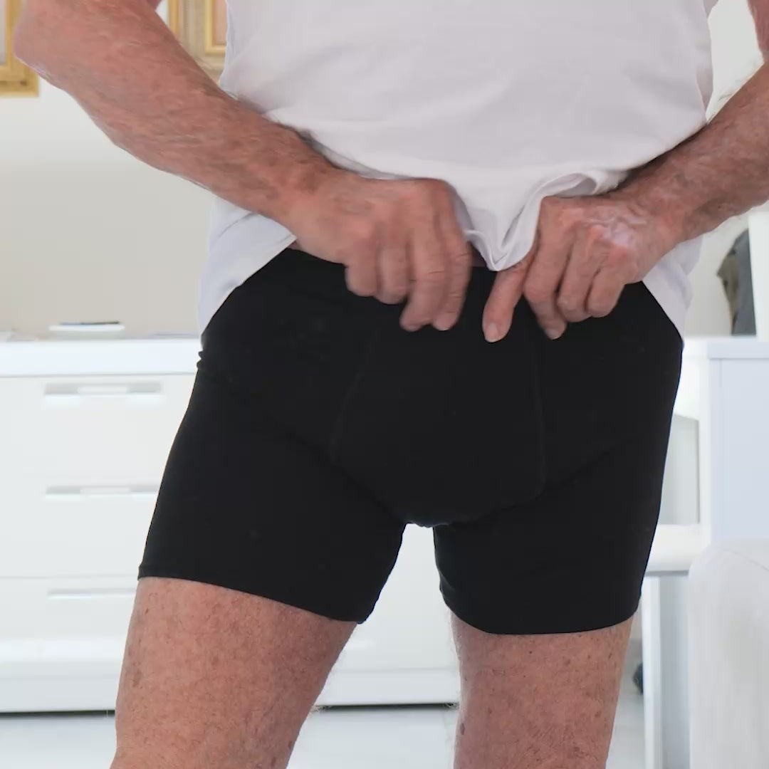 Adult man trying on Orykas absorbent boxer briefs in a bedroom, adjusting the waistband to check comfort and support, demonstrating discreet and washable underwear designed for light to moderate bladder leaks.